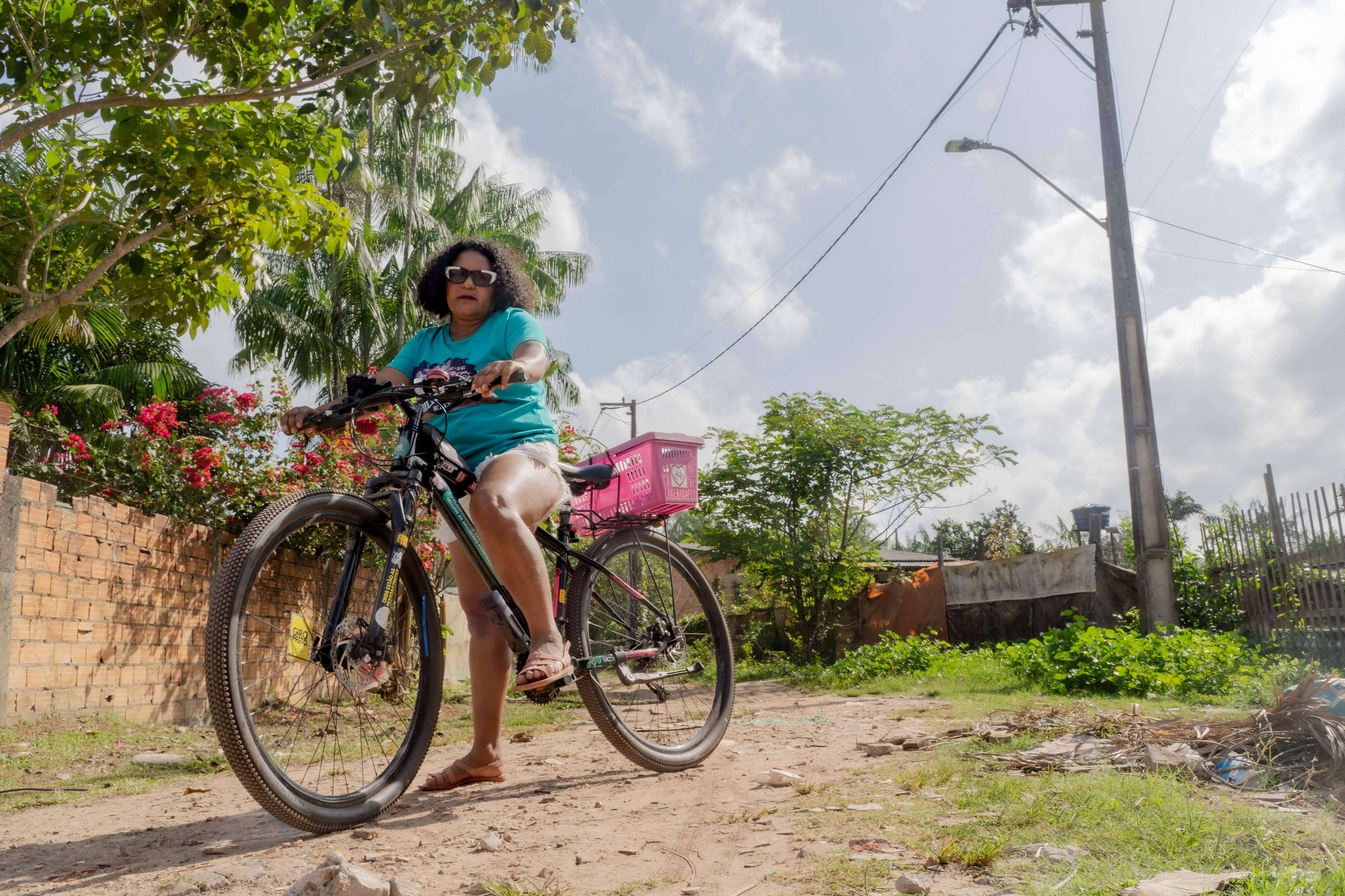 A fotografia captura uma mulher de pele escura, com cabelo cacheado escuro e óculos escuros, pedalando uma bicicleta em uma estrada de terra. Ela veste uma camiseta turquesa e shorts jeans. Na garupa da bicicleta, há uma cesta rosa. O ambiente é rural ou de periferia, com vegetação, muros de tijolos e um poste de energia elétrica ao fundo. A luz do sol ilumina a cena.