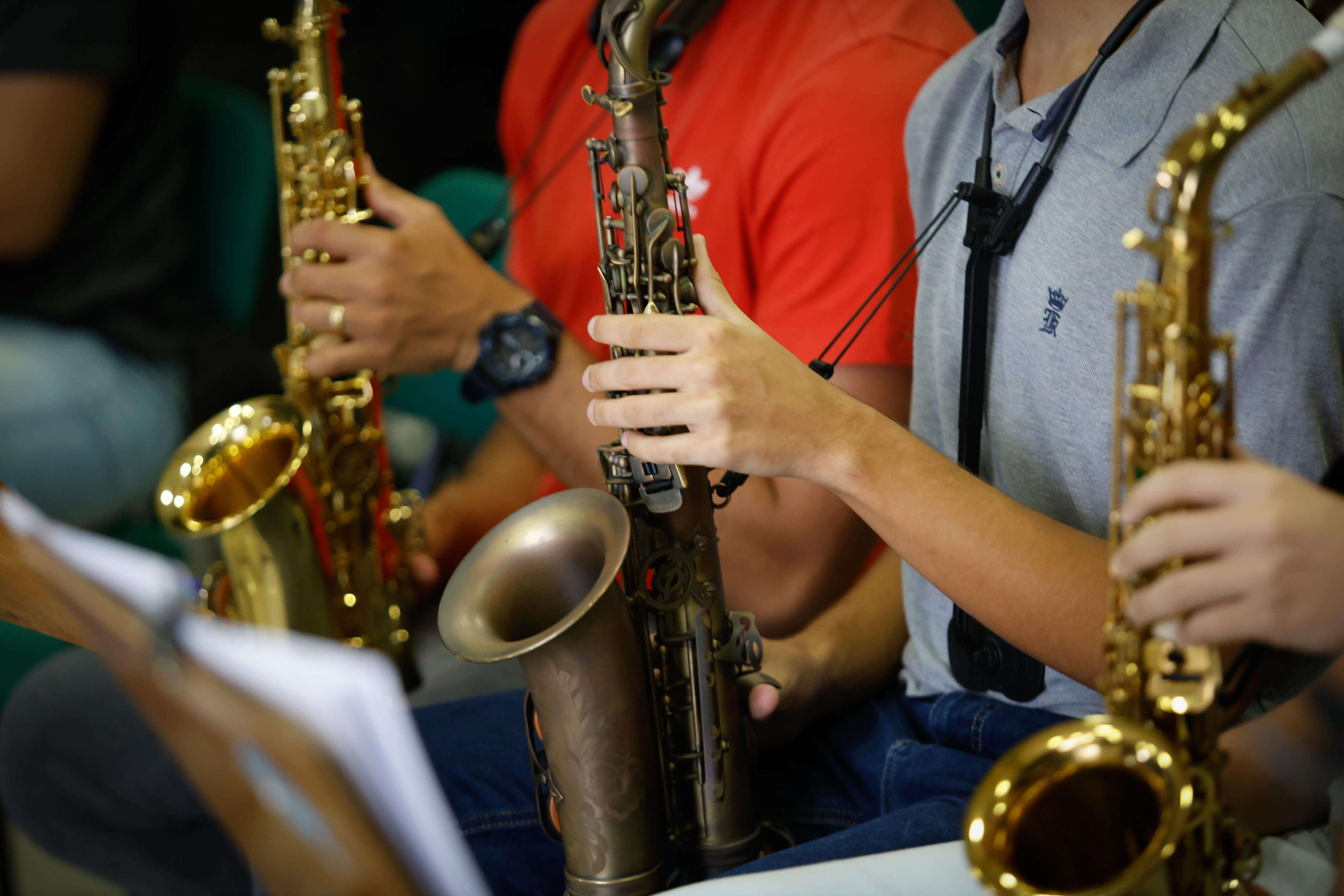 A foto é um close-up das mãos de pessoas tocando saxofones. Três músicos são visíveis, com seus instrumentos dourados e prateados. No centro, um par de mãos segura um saxofone de tom mais escuro. As unhas são curtas e limpas. À esquerda, um saxofone dourado é visível, com parte do corpo do músico em segundo plano, que veste uma camiseta vermelha. À direita, outro saxofone dourado é visível, com o braço do músico em primeiro plano, vestindo uma camiseta polo cinza. A foto foca na interação das mãos com os instrumentos, transmitindo a ideia de uma aula ou ensaio musical. Há partituras em um suporte no canto inferior esquerdo.